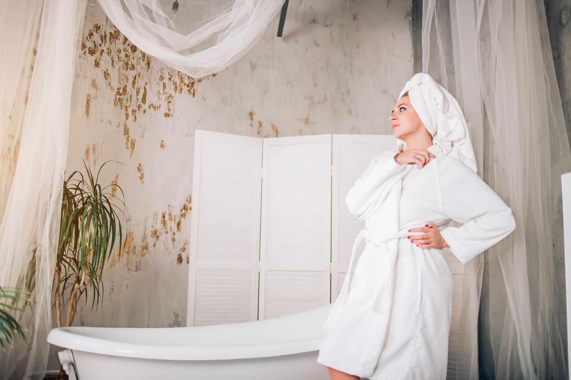 Young beautiful spa woman wearing white bathrobe and towel on head standing near bathtub in modern bathroom with loft interior looking to side thinking, making plans witn serious expression